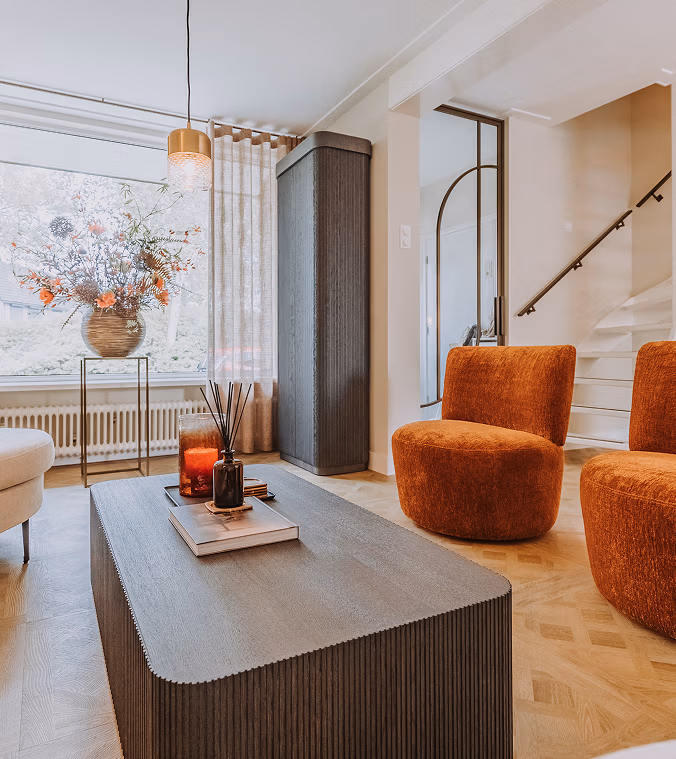 Een moderne woonkamer met twee oranje accentstoelen, een geribbelde donkere salontafel met kaarsen en boeken, een hoge grijze kast, een groot raam met transparante gordijnen en een vaas met bloemen op de vensterbank.