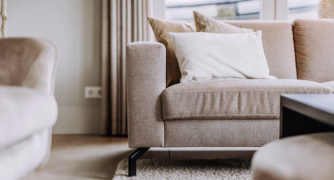 Close-up van een beige bank met een wit kussen en een kussen met structuur, geplaatst op een lichtgekleurd tapijt in een lichte woonkamer met grote ramen en gordijnen in neutrale kleuren.