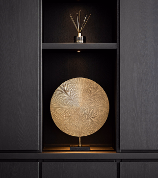 Op een zwarte plank staat een ronde, decoratieve, goudkleurige schijf op een standaard. Deze wordt verlicht door een spotje. Op de plank erboven is een geurstokjesverspreider geplaatst.