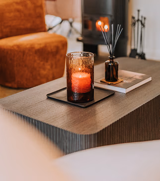 Een glazen kaarsenhouder met een brandende rode kaars staat op een donkere houten salontafel naast een boek en een geurstokjes. Op de achtergrond staan een gezellige stoel en een open haard.