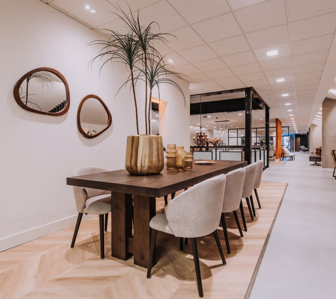 Een moderne eethoek met een donkere houten tafel, zes lichte gestoffeerde stoelen, een grote potplant als middelpunt, twee onregelmatige spiegels aan de muur en een ruim, stijlvol interieur daarachter.