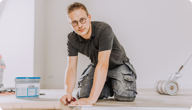 Een man met een bril en werkkleding knielt op de vloer en werkt aan een klusje in huis. Op de achtergrond zijn een container en vloerschuurapparatuur zichtbaar.