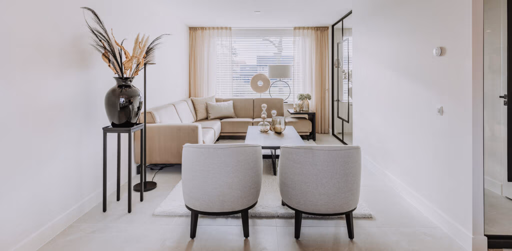 Een moderne, neutrale woonkamer met een beige hoekbank, twee grijze fauteuils, een zwarte salontafel, een grote vaas met gedroogde planten en een raam met witte jaloezieën die natuurlijk licht binnenlaten.