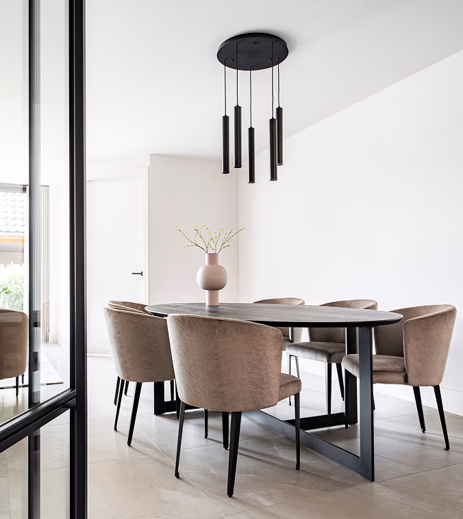 Een moderne eetkamer met een zwarte ovale tafel, zes beige gestoffeerde stoelen, een minimalistische hanglamp en een roze vaas met delicate takken op tafel. De ruimte heeft helder natuurlijk licht en neutrale tinten.