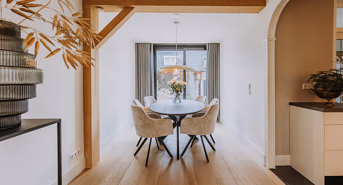 Moderne eetkamer met ronde tafel, zes gestoffeerde stoelen, vaas met bloemen, houten vloer, grote ramen met grijze gordijnen en zacht natuurlijk licht.