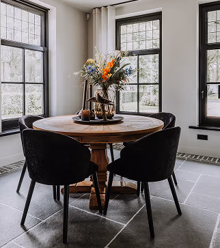 Een ronde houten eettafel met een centraal element en vijf zwarte stoelen staat in een lichte kamer met grote ramen, een grijze stenen tegelvloer en witte muren.