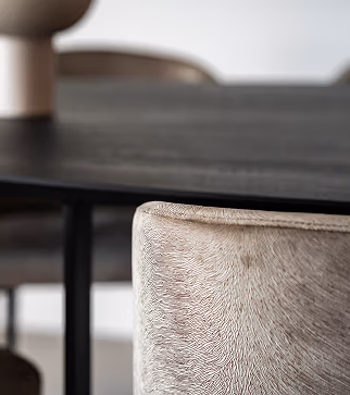 Close-up van een moderne eettafel met een lichtgekleurde stoel met textuur op de voorgrond en een donker tafelblad. De achtergrond is zacht vervaagd, waardoor de stof en de rand van de stoel goed zichtbaar zijn.