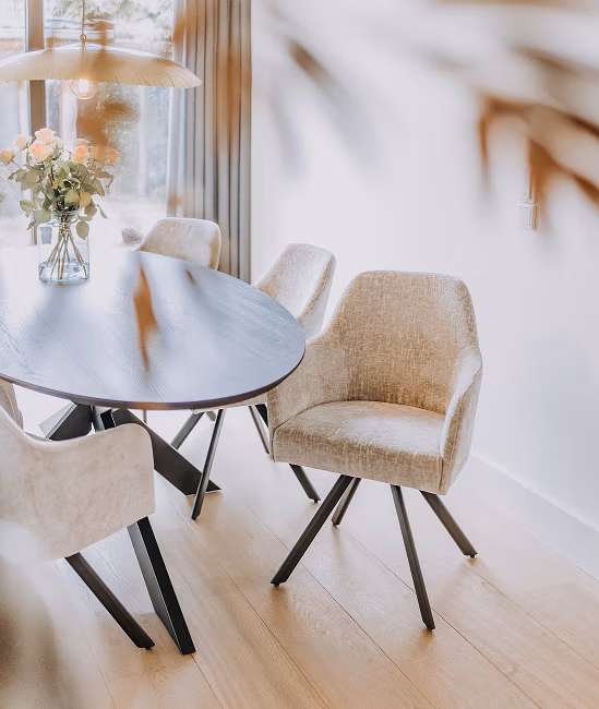 Een moderne eethoek met een donkere ovale tafel, beige gestoffeerde stoelen en een vaas met roze rozen op tafel, verlicht door natuurlijk licht via de grote ramen.