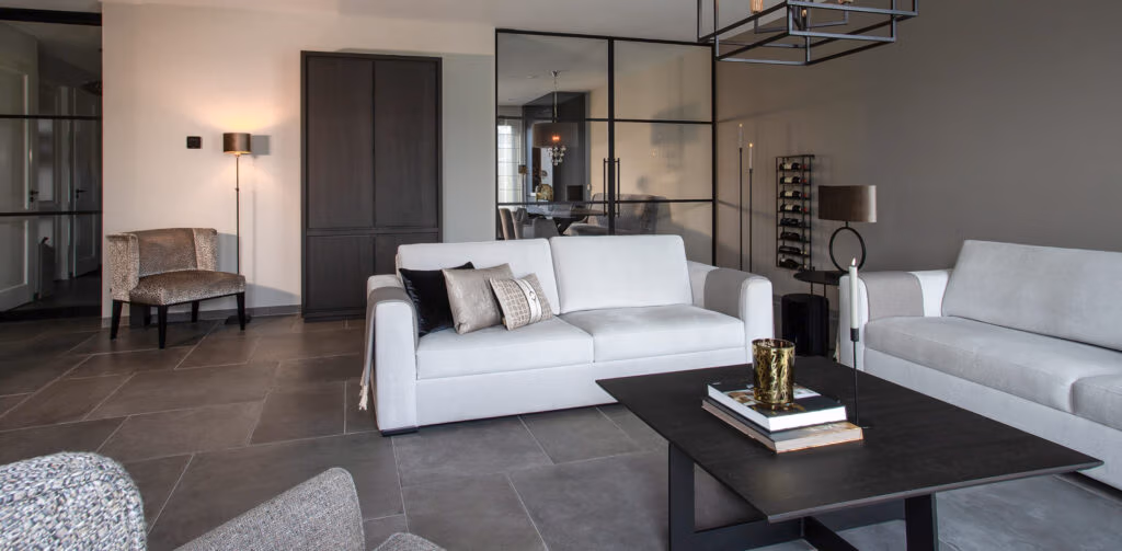 Een moderne woonkamer met grijze banken, een zwarte salontafel met boeken en decoratie, een donkere kast, glazen schuifdeuren en neutrale muren en vloeren. In de hoek staat een stoel bij een wandlamp.