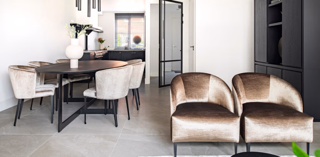 Moderne woon- en eetkamer in neutrale tinten, met twee fluwelen stoelen, een ronde eettafel met zes stoelen, een witte vaas met bloemen en een zwarte kast. Zonlicht stroomt door het raam op de achtergrond.