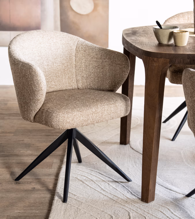 Een moderne eetkamerstoel met lichtbruine bekleding en zwarte metalen poten staat naast een houten eettafel. Een uitnodigende opstelling geïnspireerd op Japandi-meubels, compleet met beige kommen en kopjes op een lichtgekleurd tapijt op een houten vloer.