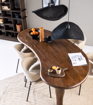 Een niervormige houten eettafel met beige stoelen, zwarte hanglampen erboven en accenten van Japandi-meubels, met een dienblad met een kopje en snacks, kommen met sinaasappels, decoratieve vazen en een opengeslagen tijdschrift op een beige vloerkleed met textuur.