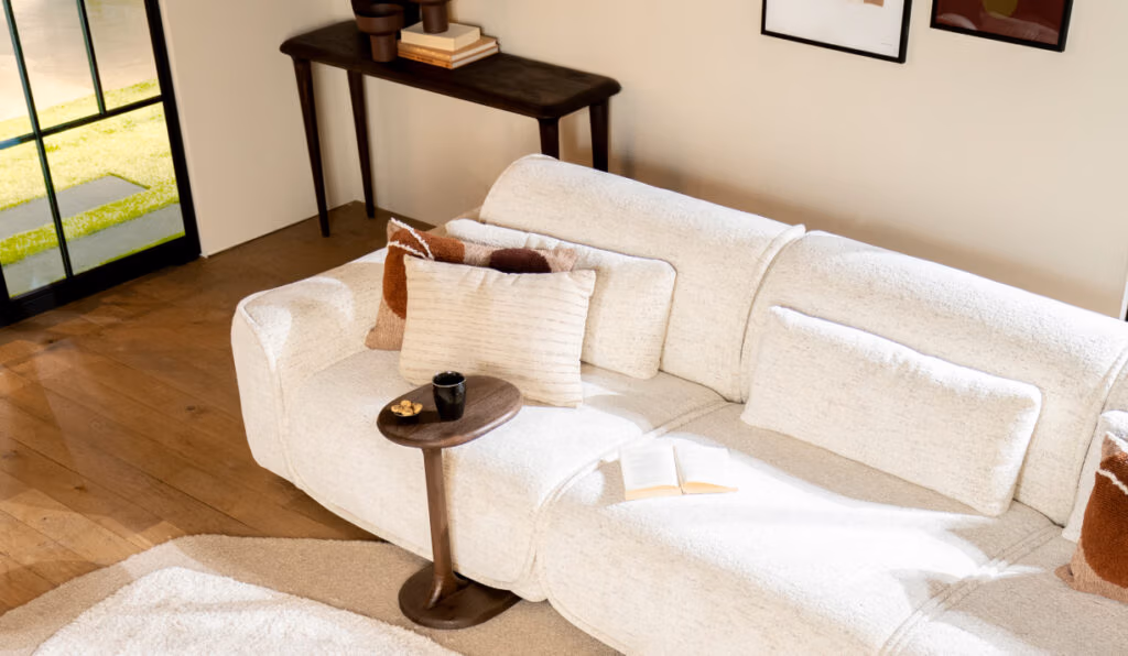 Een lichte woonkamer met Japandi-meubels: een witte bank met bruine en witte kussens, een klein houten bijzettafeltje met een kopje, snacks en een open boek. Het zonlicht stroomt door de glazen deuren naar binnen op de houten vloer.