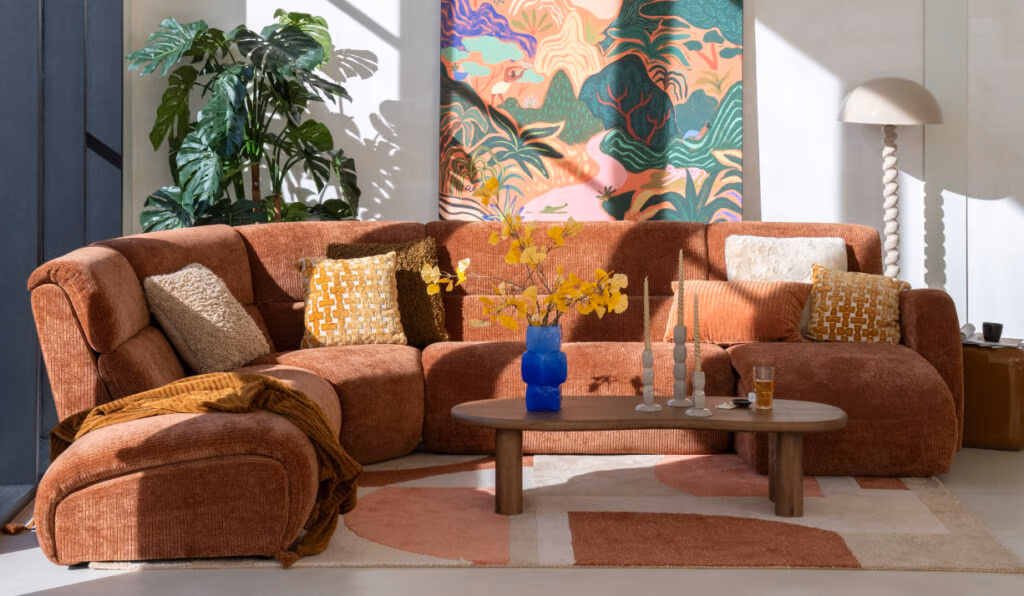 Een gezellige woonkamer met moderne meubels, waaronder een roestkleurige hoekbank, kussens met patroon, een houten salontafel met kaarsen, een blauwe vaas gevuld met gele bloemen, een bladplant en een kleurrijk abstract schilderij aan de muur.