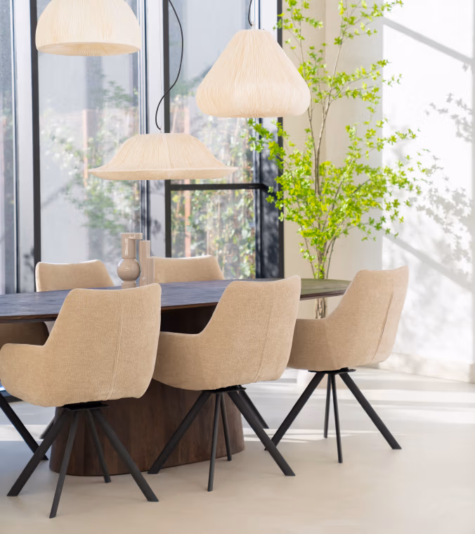 Moderne eethoek met modern meubilair, waaronder beige gestoffeerde stoelen rond een donkere ovale tafel, drie gevlochten hanglampen aan het plafond, grote ramen die de ruimte vullen met natuurlijk licht en een hoge groene plant in de hoek.