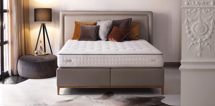 Een moderne slaapkamer met een groot bed met een gecapitonneerde matras, decoratieve kussens in verschillende kleuren, een gewatteerd hoofdeinde, een zwarte staande lamp met drie poten en sierlijke witte wandlijsten. Natuurlijk licht komt binnen via een raam.