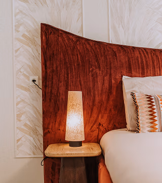 Een modern nachtkastje met een structuurlamp staat naast een bed met een hoog, donkerrood fluwelen hoofdeinde en kussens met patroon tegen een lichtgekleurde muur met subtiele bladmotieven.