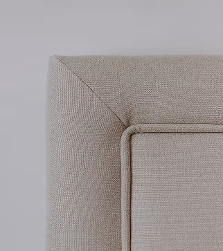 Close-up van de bovenhoek van een beige gestoffeerd hoofdbord met zichtbare stiksels en een structuurstof afwerking tegen een lichtgrijze achtergrond.