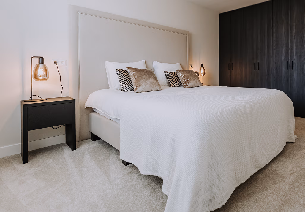 Moderne slaapkamer met een groot bed met een wit dekbed, twee decoratieve kussens, zwarte nachtkastjes met glazen lampen en een donkere inbouwkast tegen een witte muur.