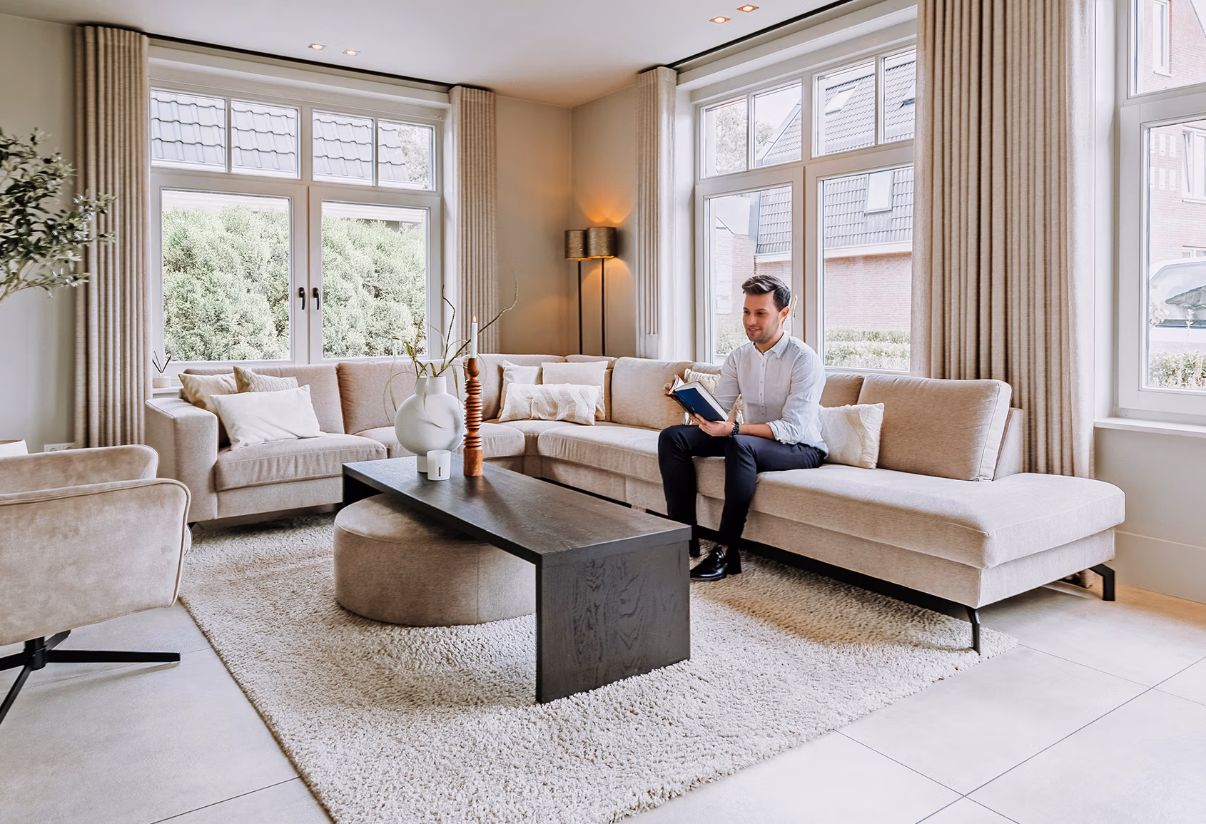 Een man zit te lezen op een beige hoekbank in een lichte, moderne woonkamer met grote ramen, een neutrale inrichting, een licht tapijt, een donkere salontafel en minimalistische decoraties.
