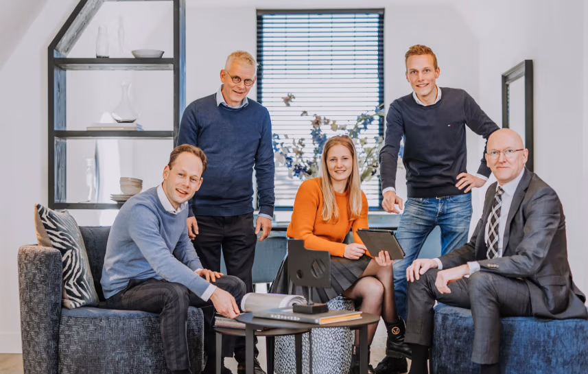 Vijf mensen in zakelijke kleding zitten en staan rond een kleine salontafel met laptops en documenten. Ze lachen naar de camera in een moderne, goed verlichte kantoorruimte.