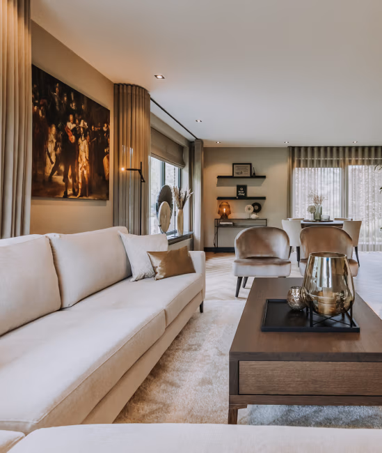 Moderne woonkamer met een beige bank, twee fauteuils, een houten salontafel met decoratieve elementen, grote ramen met transparante gordijnen en een eethoek op de achtergrond. Muurkunst en zachte verlichting zorgen voor een gezellige sfeer.