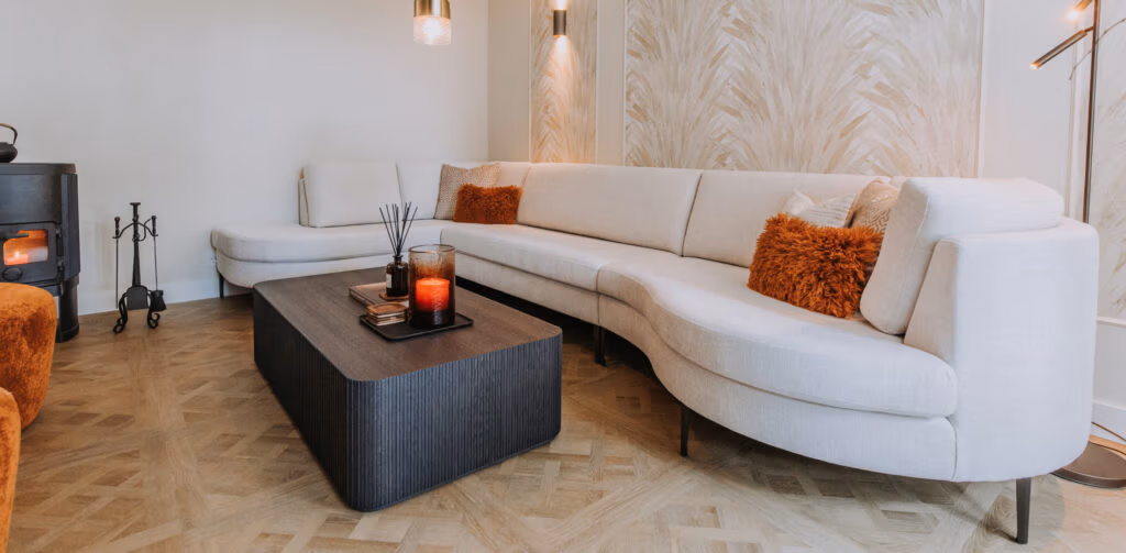 Een moderne woonkamer met een grote, gebogen witte bank, oranje kussens met textuur, een donkere, rechthoekige salontafel met kaarsen en decoratie, en een houten vloer met patroon. Lampen en zachte verlichting zorgen voor een gezellige sfeer.