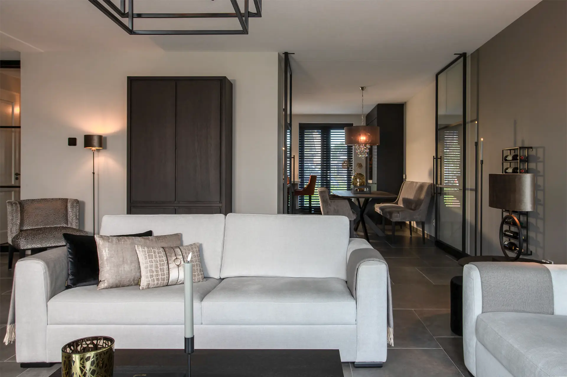 Moderne woonkamer met een lichtgrijze bank, donkere accentkussens en een salontafel ervoor. Op de achtergrond is de eethoek met een zwarte tafel en stoelen zichtbaar, met grote ramen en een eigentijdse inrichting.