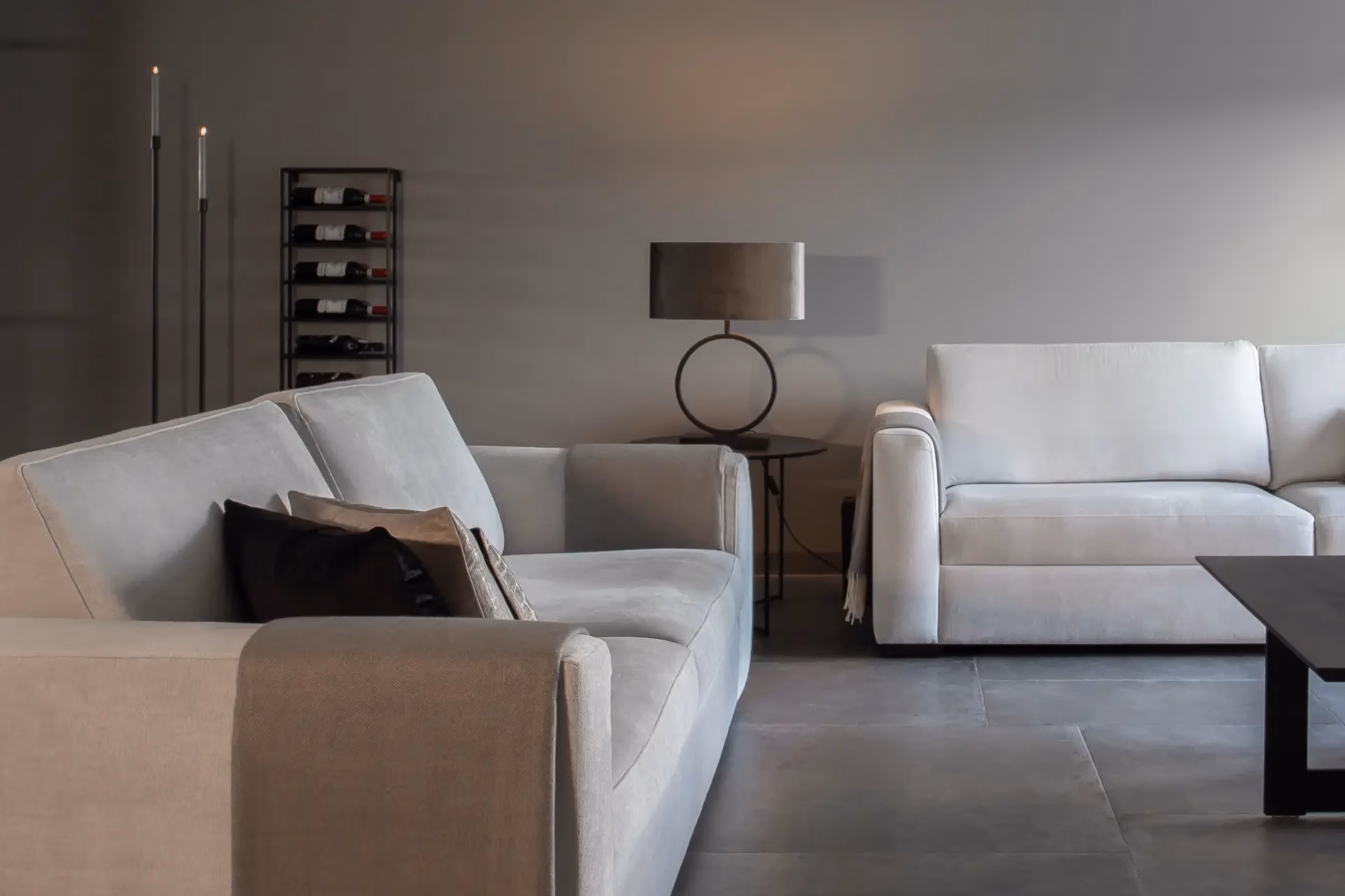 Een moderne woonkamer met twee lichtgrijze banken, een zwarte salontafel, een ronde lamp op een bijzettafel en een wijnrek tegen een grijze muur. De ruimte heeft een minimalistische en neutrale inrichting.
