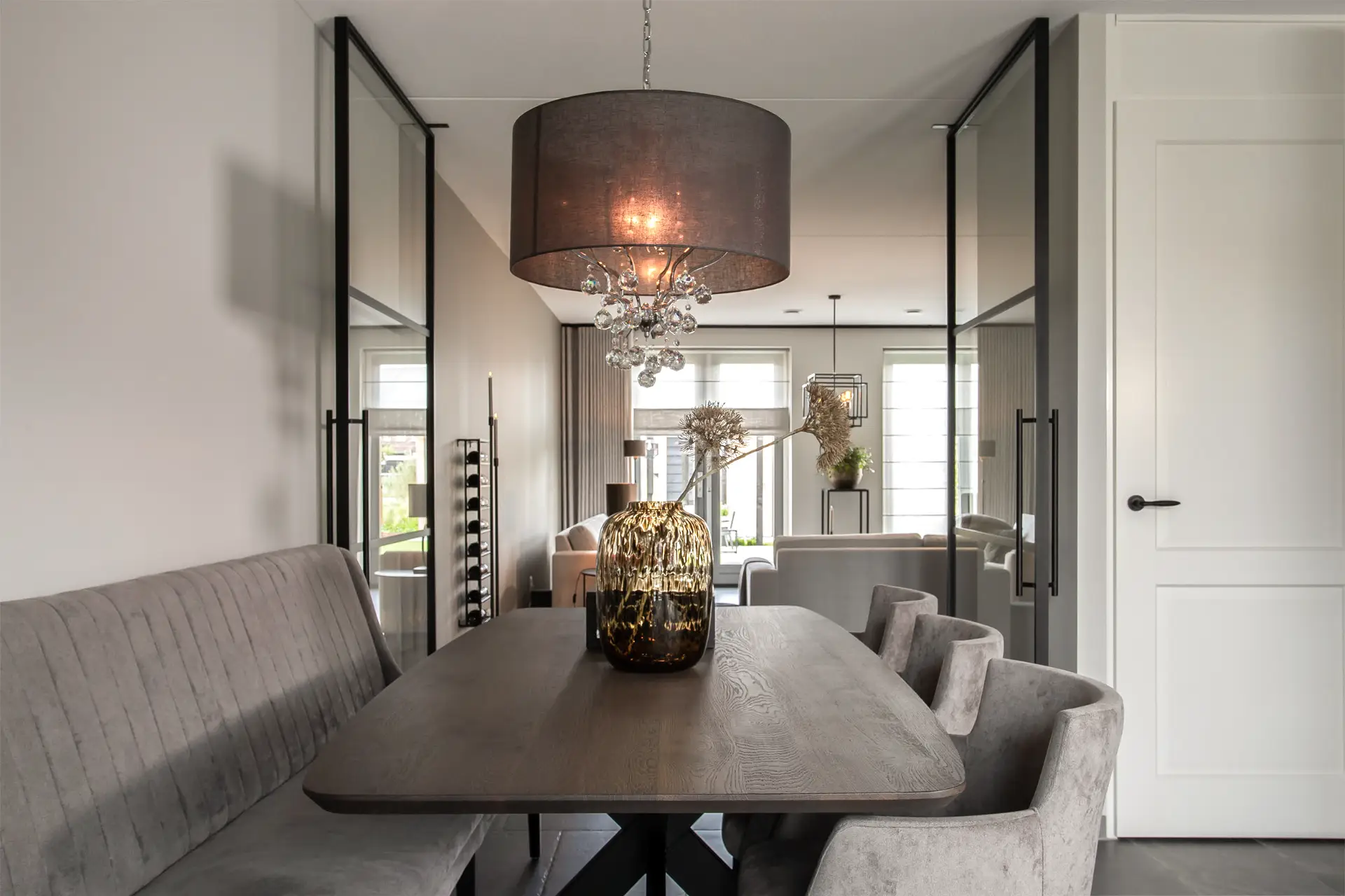 Moderne eetkamer met een donkere houten tafel, een grijze bank en stoelen, een grote decoratieve vaas als middelpunt, glazen deuren en een stijlvolle kroonluchter boven de tafel.