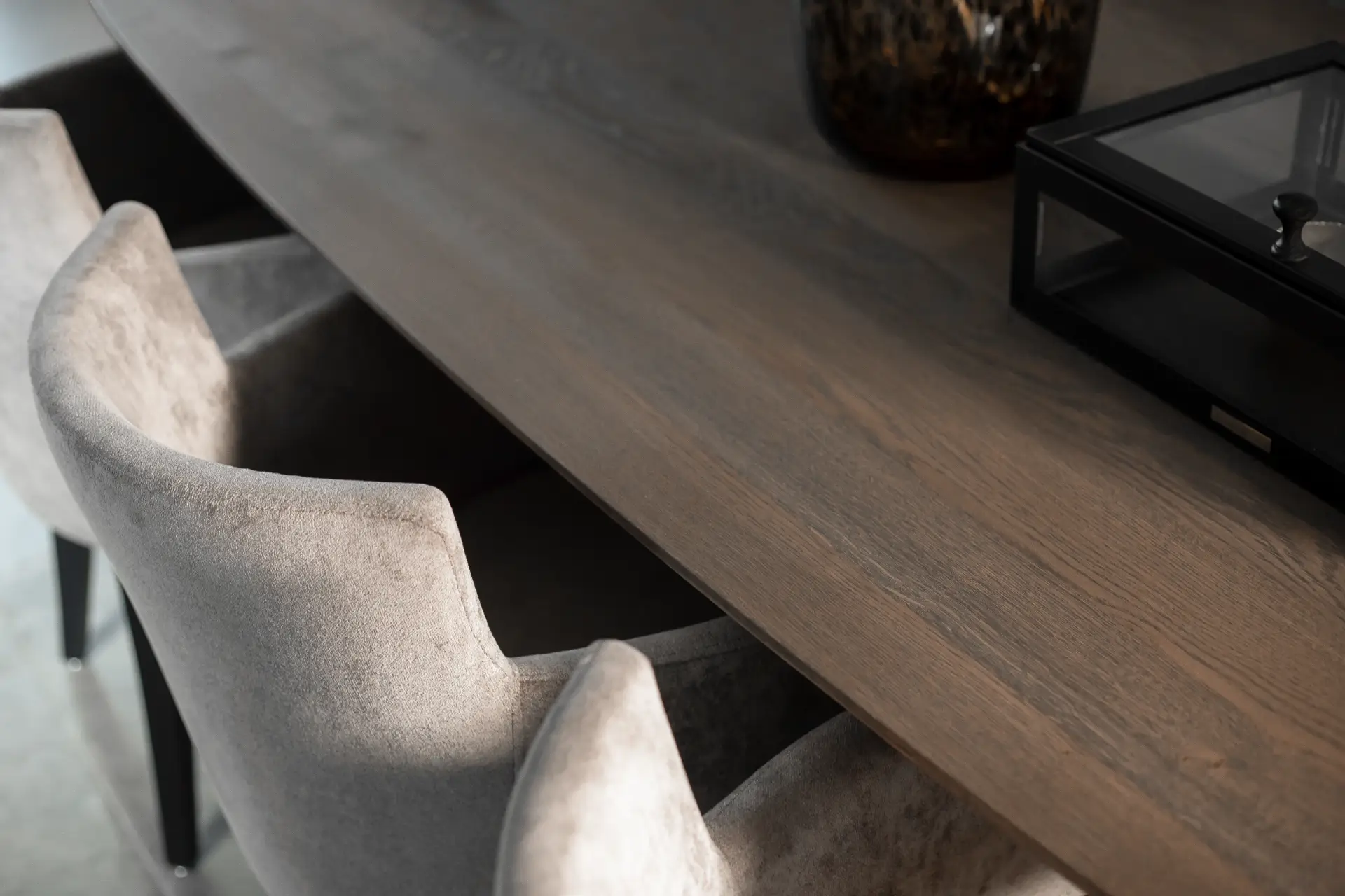 Een close-up van een houten tafel met twee beige gestoffeerde stoelen. Op het tafelblad zijn een deel van een decoratief object en een glazen doos te zien.