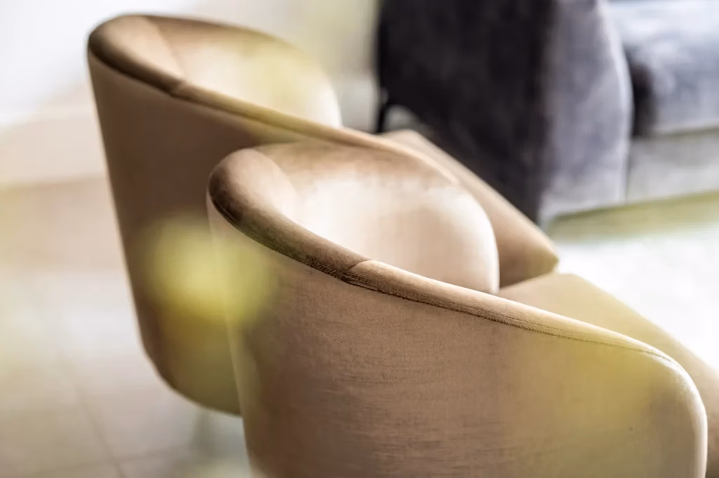 Twee beige fluwelen fauteuils met gebogen rugleuningen staan naast elkaar op een lichte vloer. Op de achtergrond is een donkergrijze bank zichtbaar. De verlichting is zacht en creëert een gezellige en uitnodigende sfeer.