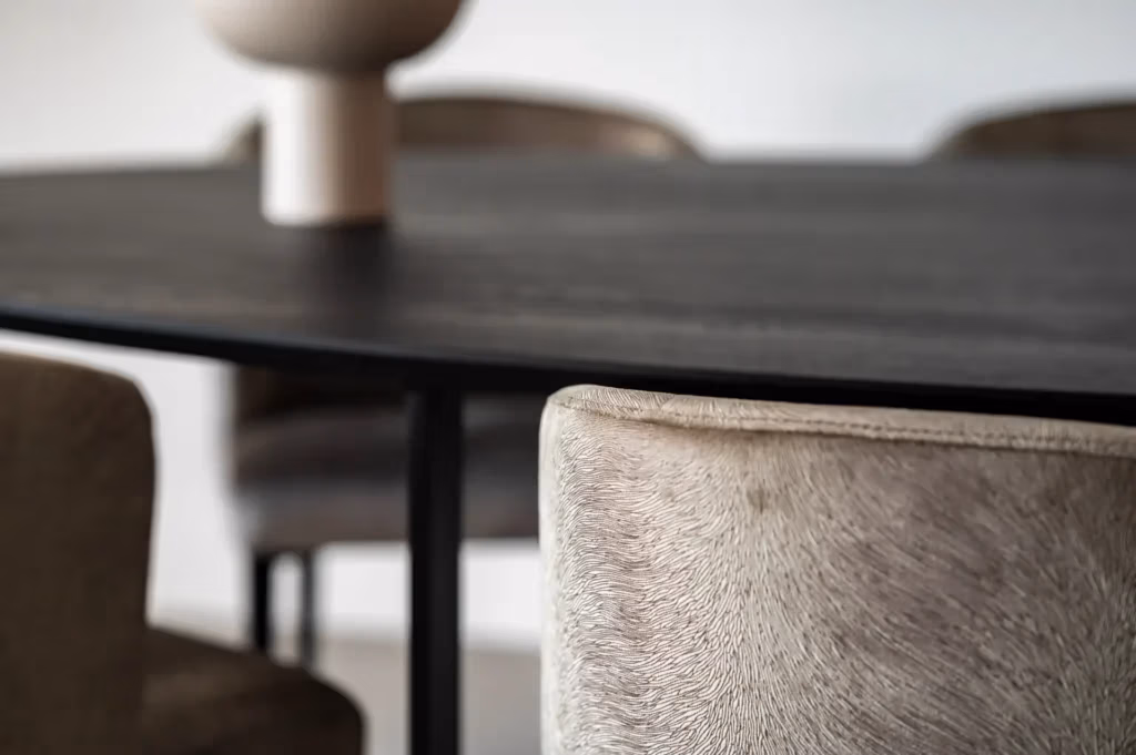 Close-up van een moderne eettafel met een donker houten blad, omringd door beige gestoffeerde stoelen. Een decoratieve vaas staat op het tafelblad. De setting heeft een minimalistische, eigentijdse stijl.