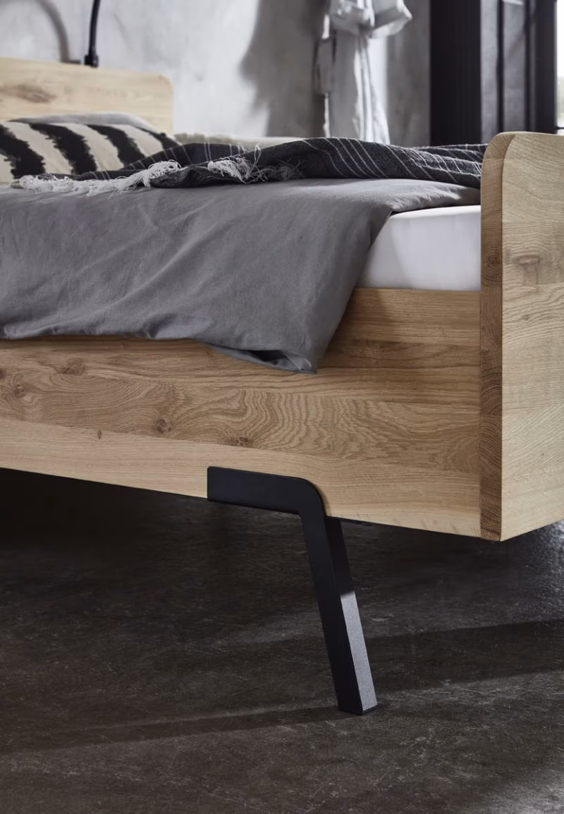 Close-up van een modern houten bedframe met schuine, zwarte metalen poten, grijs beddengoed en een zwart-wit gestreepte deken, op een donkere betonnen vloer in een minimalistische slaapkamer.