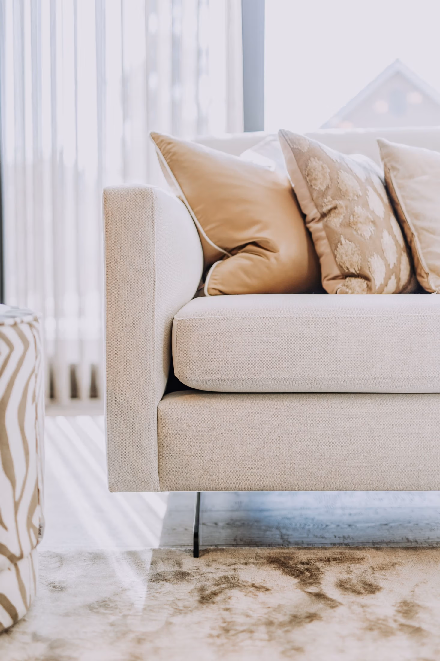 Een close-up van een moderne beige bank met zachte kussens, waaronder een met een patroon, in een lichte woonkamer. Zonlicht stroomt door de transparante gordijnen en verlicht de zachte texturen en neutrale tinten.