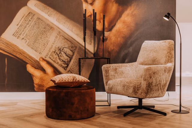Een gezellig leeshoekje met een beige fauteuil, een ronde bruine voetenbank met een kussen met patroon, een zwarte staande lamp en een bijzettafeltje, met op de achtergrond een muurschildering van een persoon die een oud boek leest.