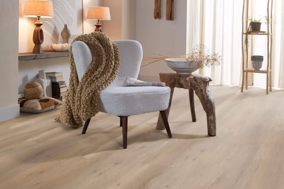 Een gezellige woonkamer met een lichtgekleurde fauteuil met een grof gebreide deken, een rustiek houten bijzettafeltje, een schaal met gedroogde planten, zachte verlichting en neutrale decoratie op planken. Door het hele huis ligt een lichte houten vloer.