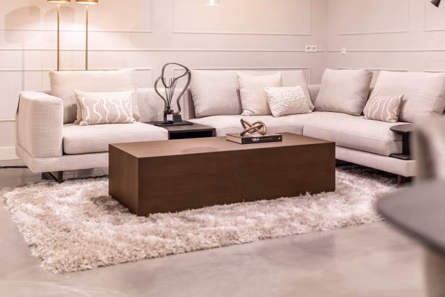 Een moderne woonkamer met een crèmekleurige hoekbank, kussens met en zonder patroon, een houten salontafel, abstracte decoratie en een zacht wit vloerkleed op een gepolijste vloer in een ruimte met neutrale tinten.