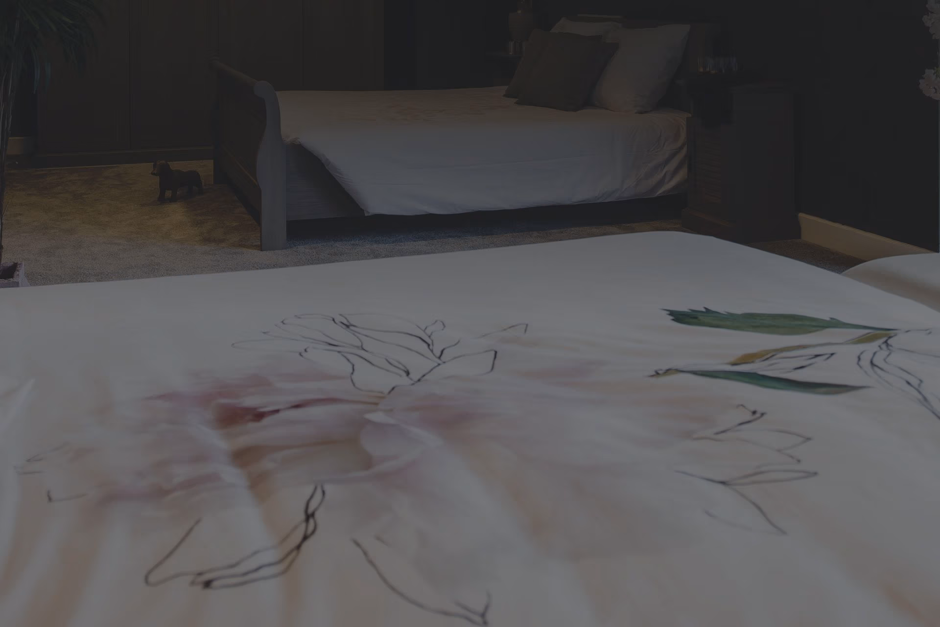 Een zacht verlichte slaapkamer met twee bedden. Op de voorgrond is een close-up van een dekbed met bloemenmotief te zien, terwijl op de achtergrond een ander netjes opgemaakt bed en wat meubels te zien zijn.