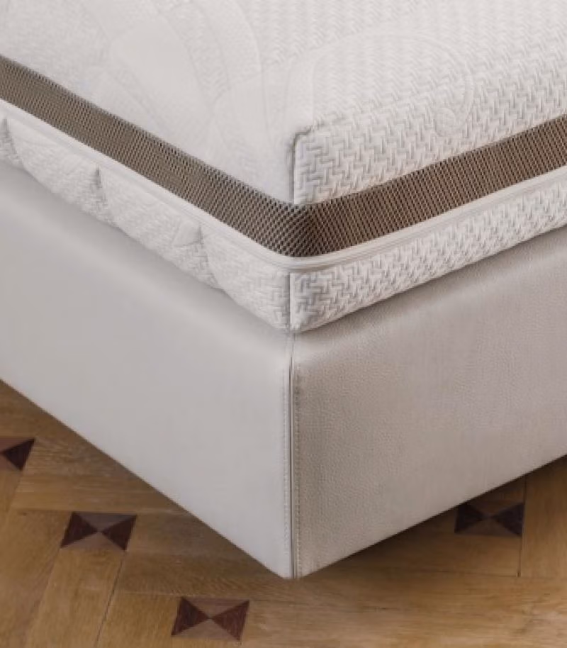 Close-up van een matras met een witte hoes met structuur en een ademend zijpaneel van mesh, rustend op een lichtgekleurd bedframe met een houten vloer met een geometrisch patroon.