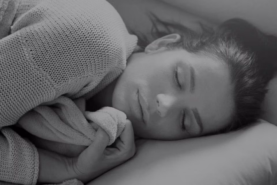 Een vrouw met donker haar slaapt vredig op haar zij, met een deken in haar hand en haar hoofd rustend op een kussen. Ze draagt een gebreide trui en oogt kalm en ontspannen.