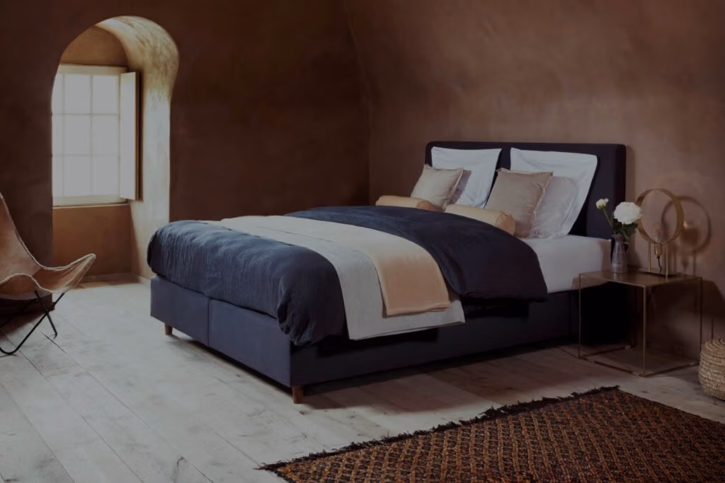 Een gezellige slaapkamer met aardse bruine muren, een groot bed met marineblauw en beige beddengoed, witte kussens, een nachtkastje met een lamp en bloemen, een vlinderstoel, een raam en een tapijt met patroon op een lichte houten vloer.