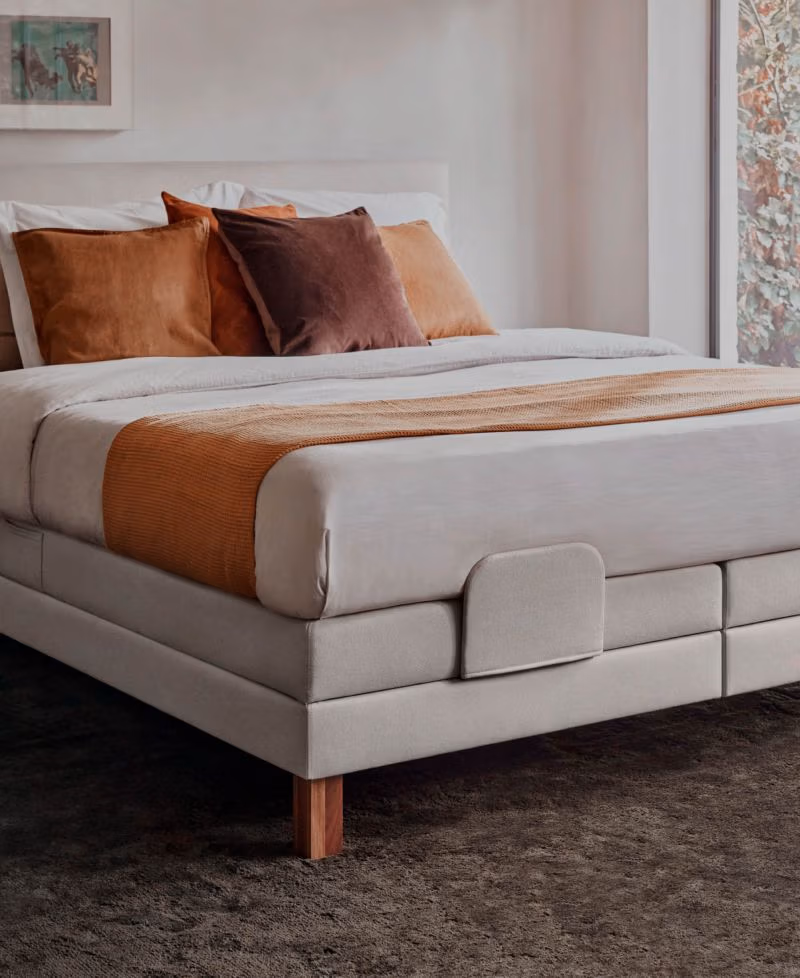 Een netjes opgemaakt bed met een beige gestoffeerd frame, wit beddengoed, oranje en bruine kussens en een bijpassend oranje sprei over het voeteneind, staat op een donker tapijt bij een groot raam met uitzicht op het groene landschap.