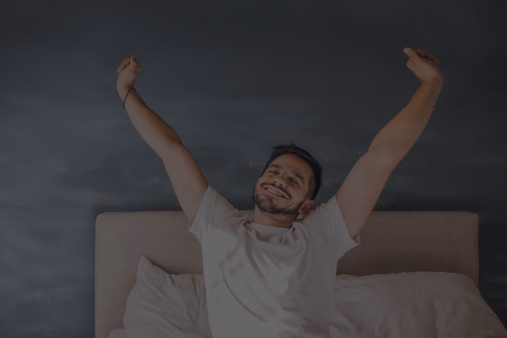 Een man zit op een bed, strekt zijn armen omhoog en glimlacht. Hij ziet er ontspannen en tevreden uit, met een donkere muur en kussens op de achtergrond.