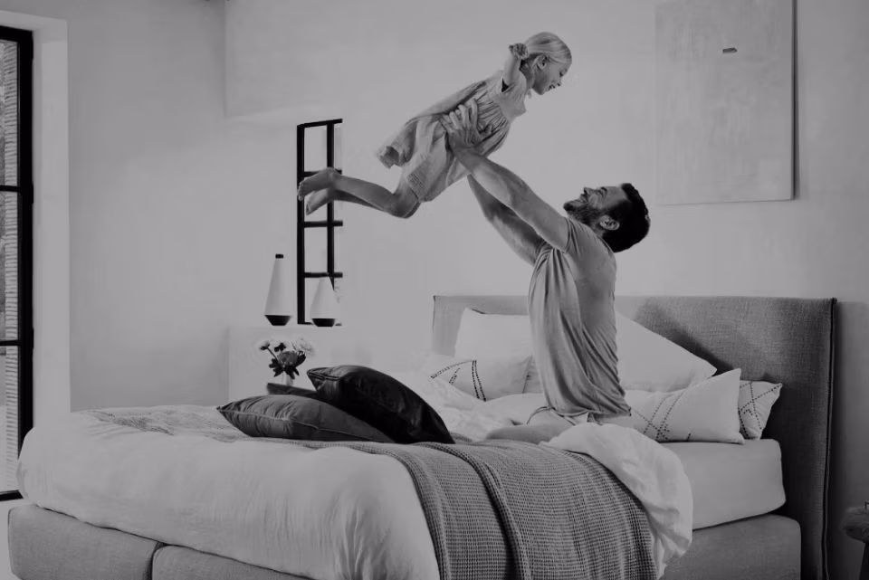 Een man op een bed tilt speels een jong kind de lucht in. De kamer is licht en gezellig, met kussens en een deken op het bed en een vaas met bloemen op het nachtkastje. Beiden lijken gelukkig en betrokken.