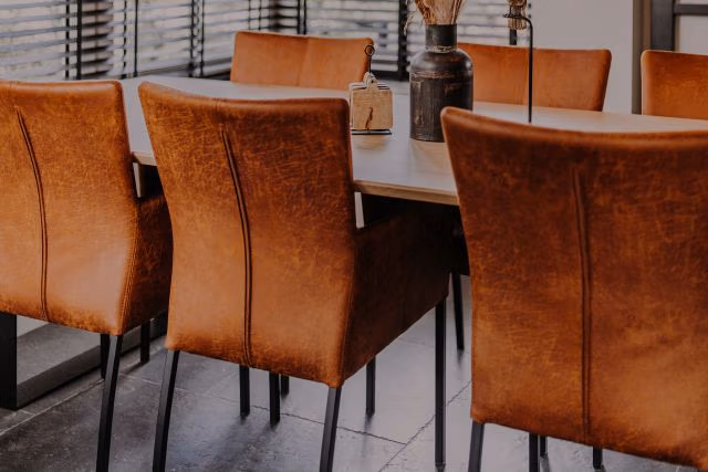 Een moderne eettafel met zes bruine leren stoelen, zwarte metalen poten en een houten tafelblad. Een donkere vaas met gedroogde bloemen staat in het midden van de tafel en jaloezieën laten natuurlijk licht binnen.