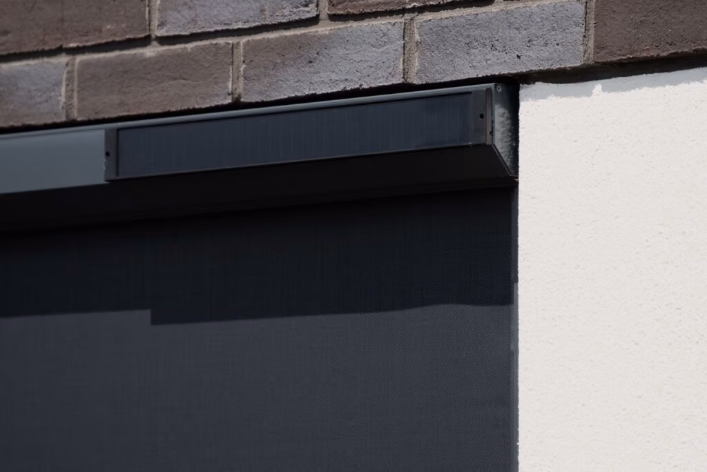 Een close-up van een modern zwart rolgordijn, gemonteerd op een witte muur onder een bruine bakstenen gevel. Het rolgordijn lijkt gedeeltelijk neergelaten, met zonlicht dat schaduwen werpt op het oppervlak.