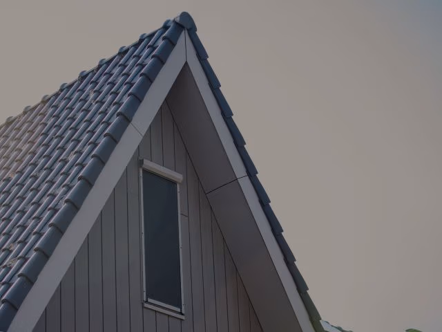 Close-up van het bovenste gedeelte van een huis met een steil, blauw pannendak en een klein verticaal raam, afstekend tegen een bleke lucht.