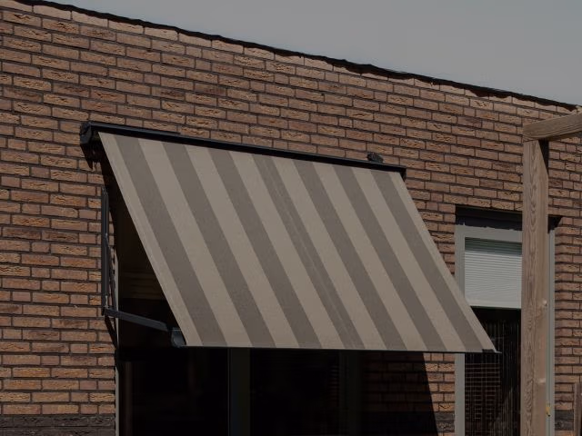 Een bruine bakstenen muur met een raam dat wordt afgedekt door een gestreept, beige en grijs uitschuifbaar zonnescherm dat schuin naar buiten loopt. Rechts is gedeeltelijk een ander raam met een gesloten wit rolgordijn zichtbaar.