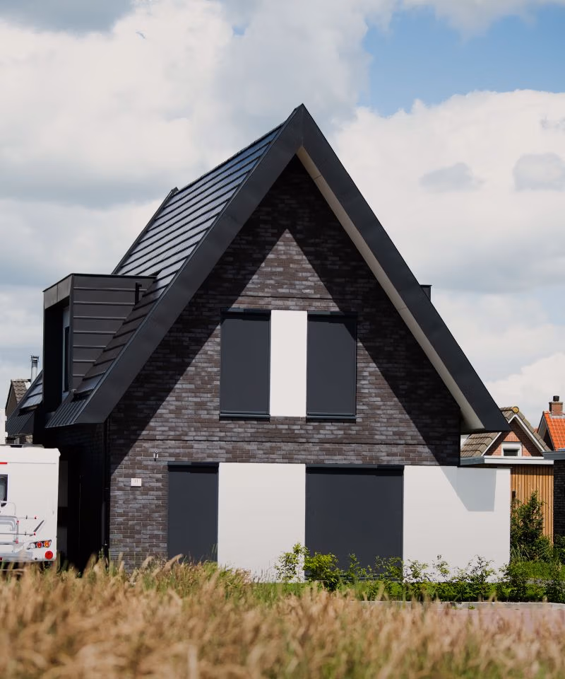 Een modern huis met donkere bakstenen muren, een groot driehoekig dak en zwart-witte luiken, omgeven door gras en een gedeeltelijk bewolkte lucht.